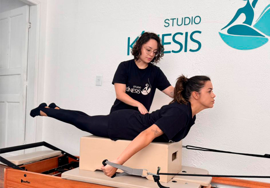 Instrutor de Pilates auxiliando aluna em exercício de alongamento em reformer em estúdio moderno
